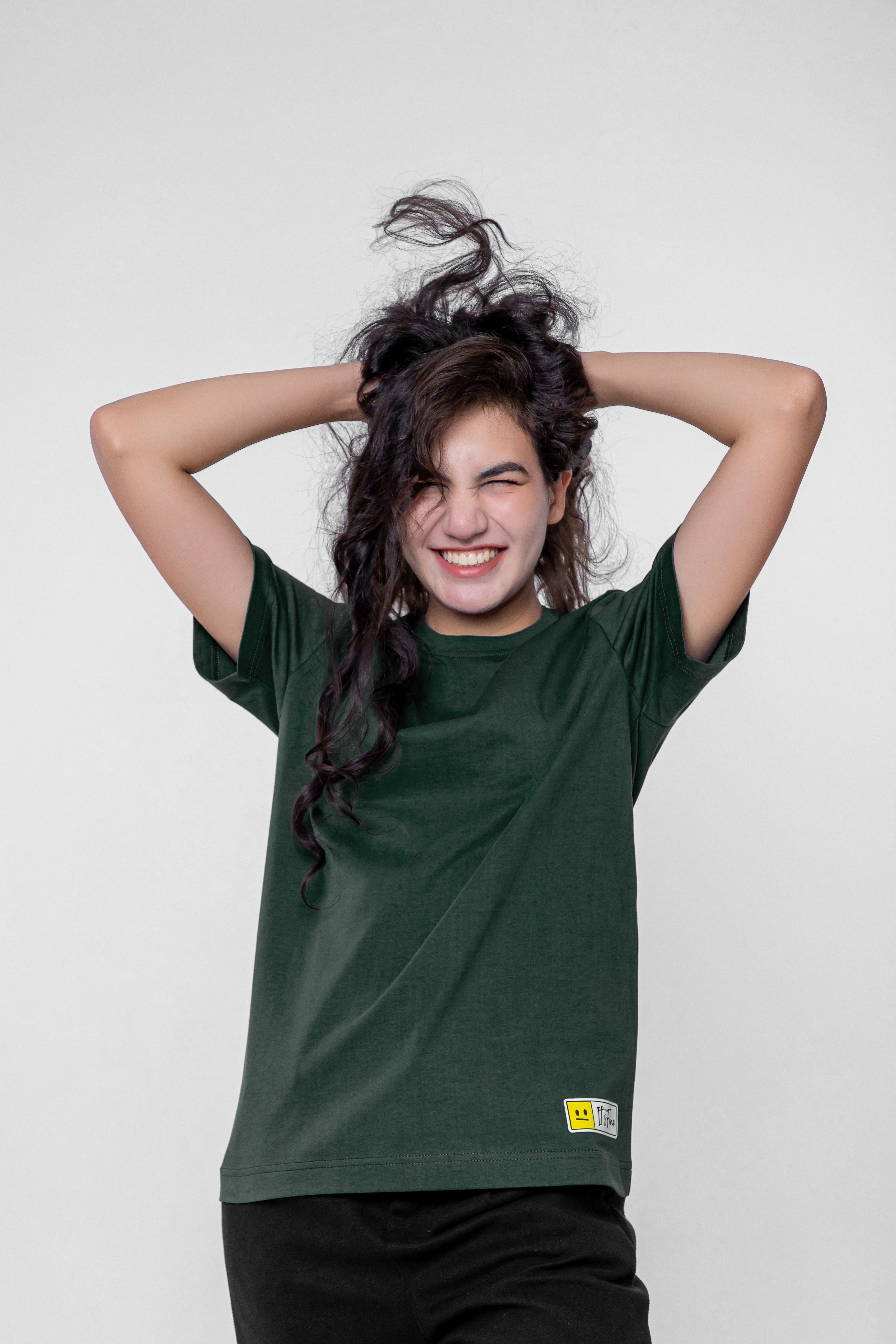 Cheerful young woman in green t-shirt smiling with hands in hair against white background.