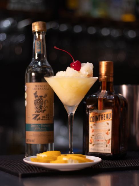 A square bottle of Cointreau next to a glass poured over ice with fresh orange peel curls on a bar counter.