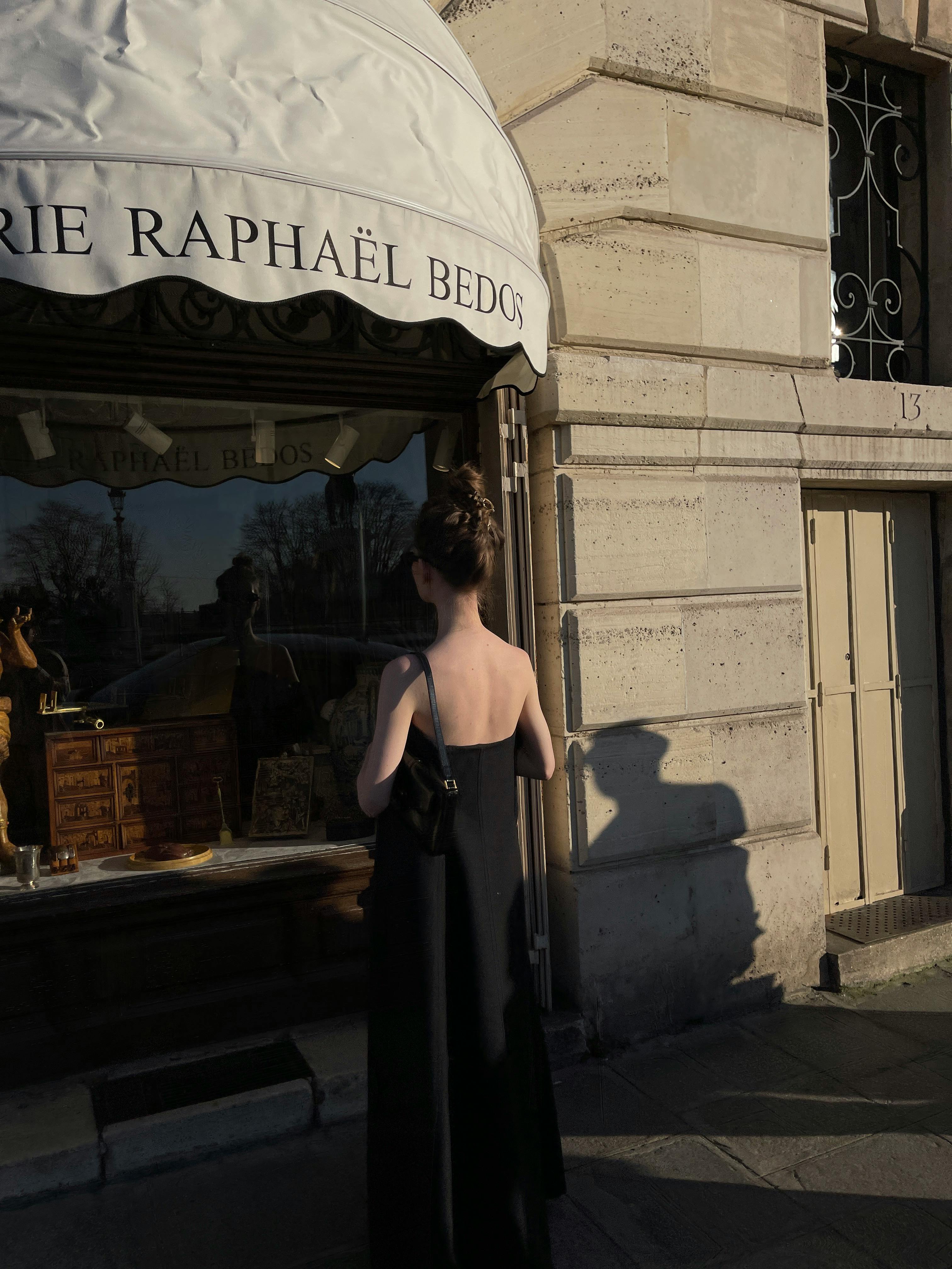 A woman in a black dress stands outside an antique store, gazing at the window display.