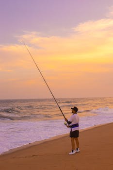 A fisherman casts his line on a sandy beach during a vibrant sunset.