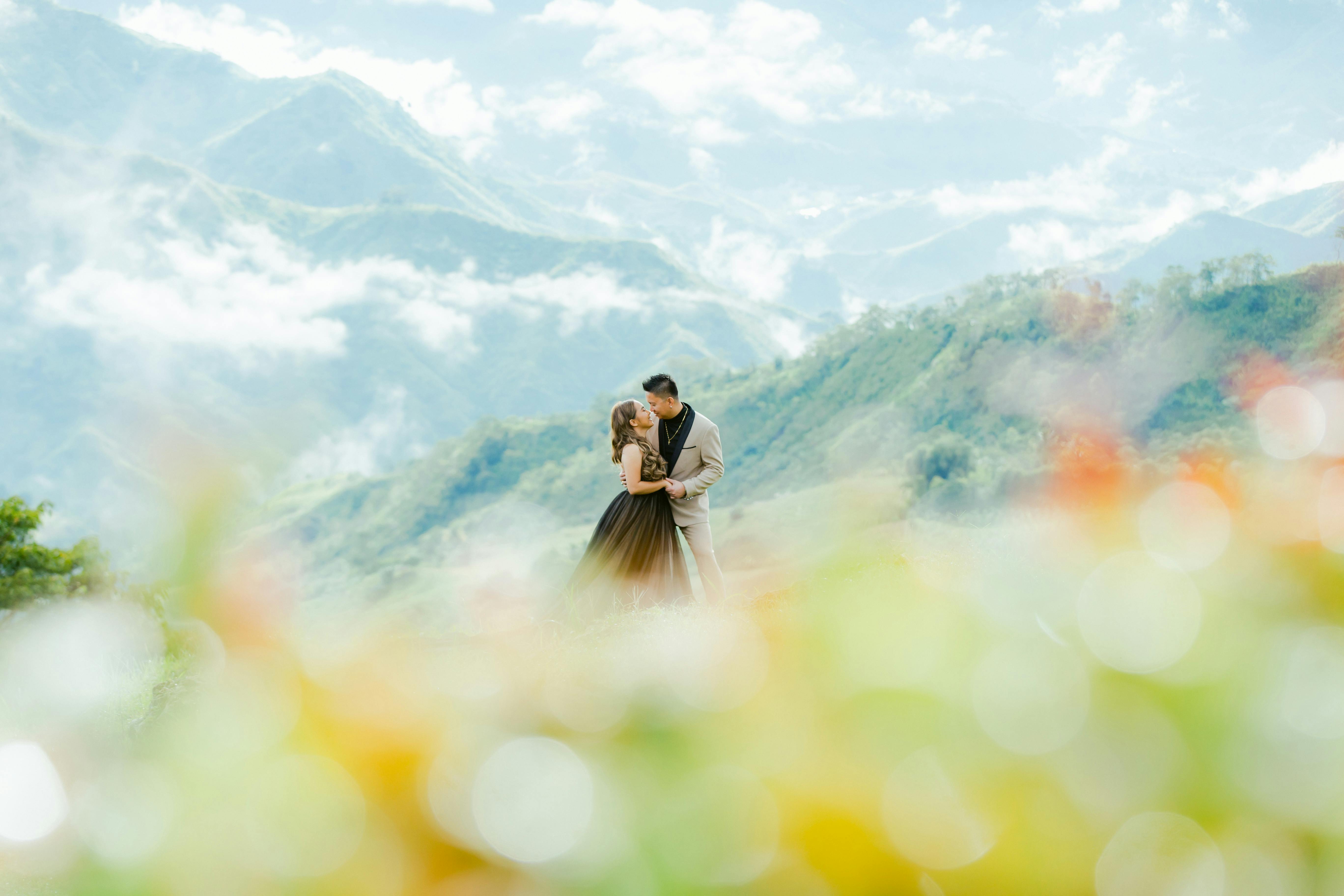 A couple shares a romantic embrace amidst a breathtaking mountain backdrop, with vibrant foreground bokeh.