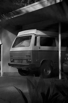 Black and white photo of a classic van parked in a shadowy carport, exuding nostalgia.