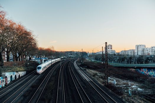 A high-speed train travels on curving tracks through an urban landscape at sunset.