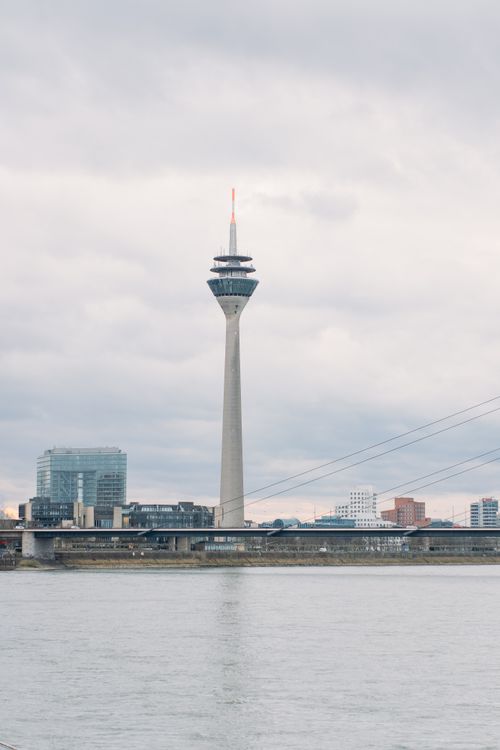Free Rhine Tower in Düsseldorf against a cloudy city skyline with the Rhine River in the foreground. Stock Photo