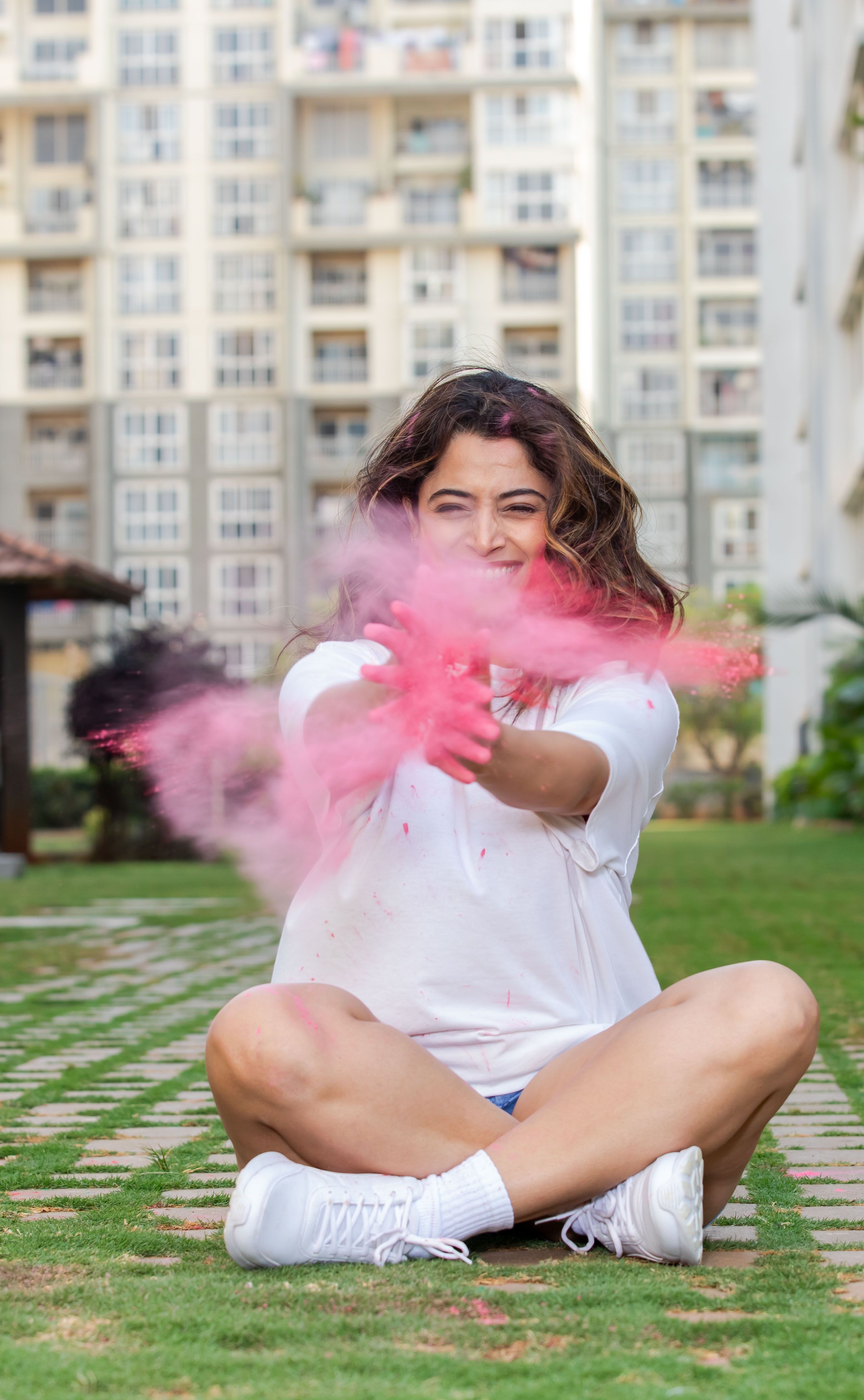 Woman in casual white clothing enjoys throwing pink colored powder outside.