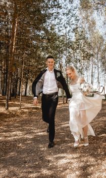 A happy couple in wedding attire runs joyfully through a sunlit forest path, capturing a candid moment.