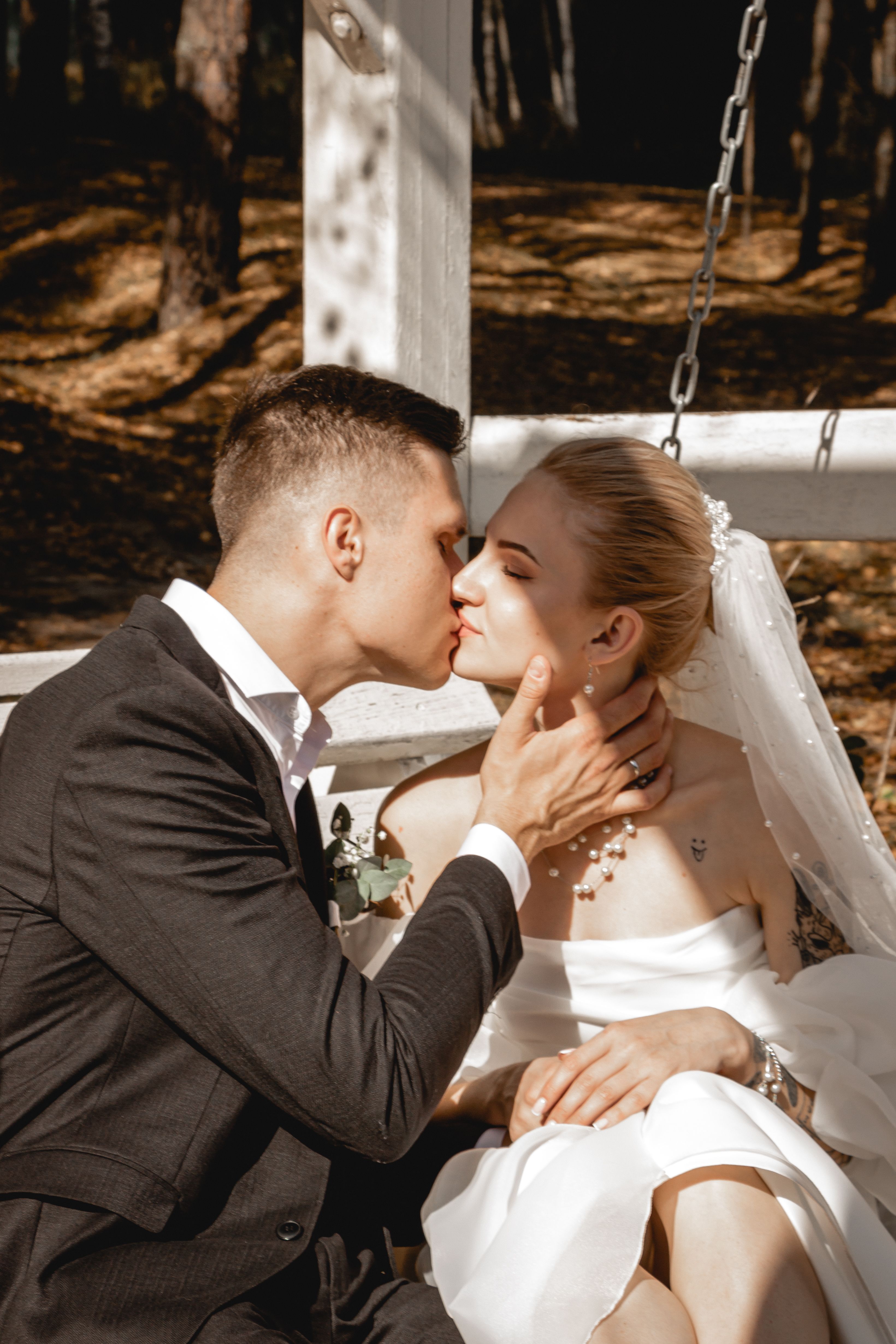 A couple shares a romantic kiss on a swing during their outdoor wedding. Sunlit scene adds warmth and love.