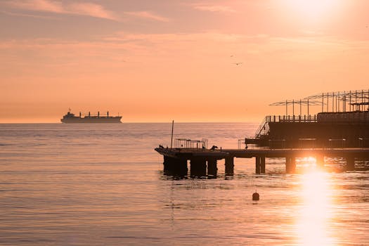 A tranquil sunrise view with a silhouette of a pier and a distant cargo ship on calm waters.