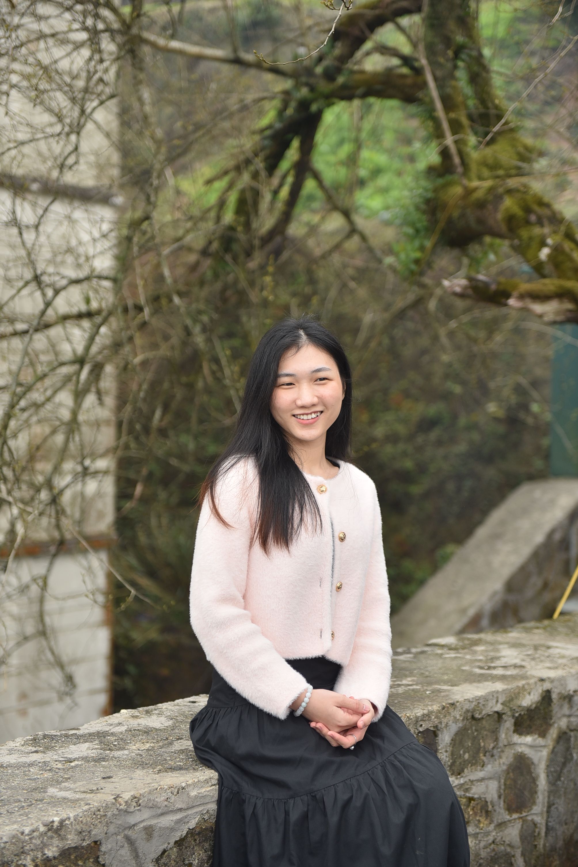 A young woman smiles sitting on a stone wall amidst lush nature.