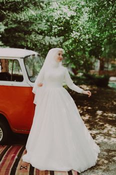 A beautiful bride in a classic white gown stands beside a retro van under lush trees.