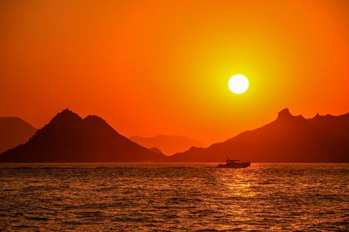 Free Silhouette of a boat sailing under a stunning sunset with mountains in the background. Stock Photo