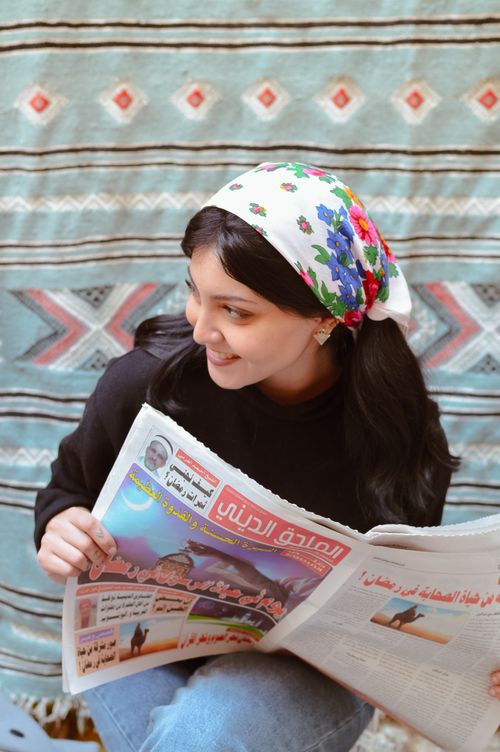 Free Smiling woman in a floral headscarf reading a newspaper indoors with vibrant tapestry background. Stock Photo