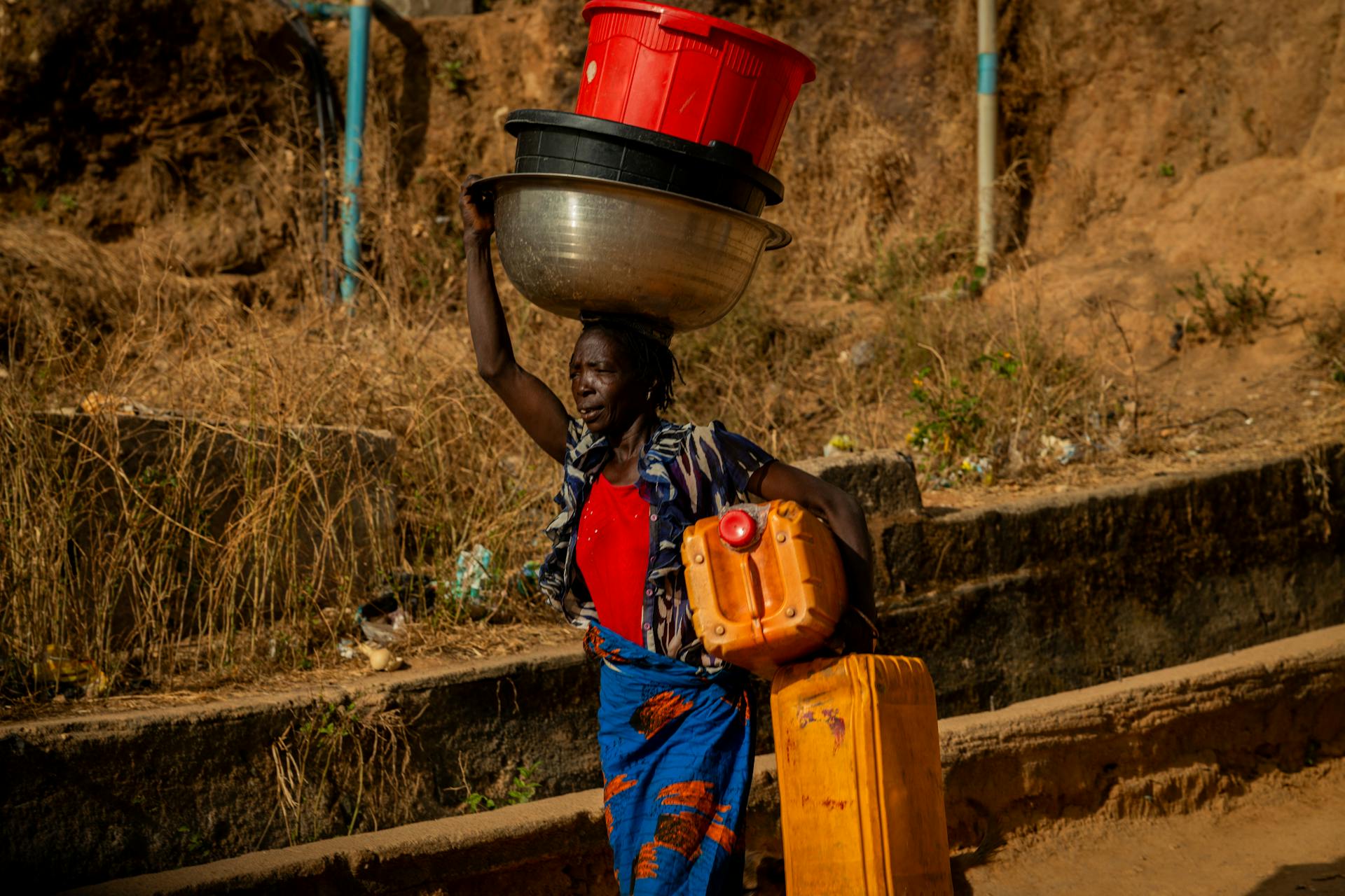 African woman balancing containers on roadside in rural area, showcasing strength and resilience.