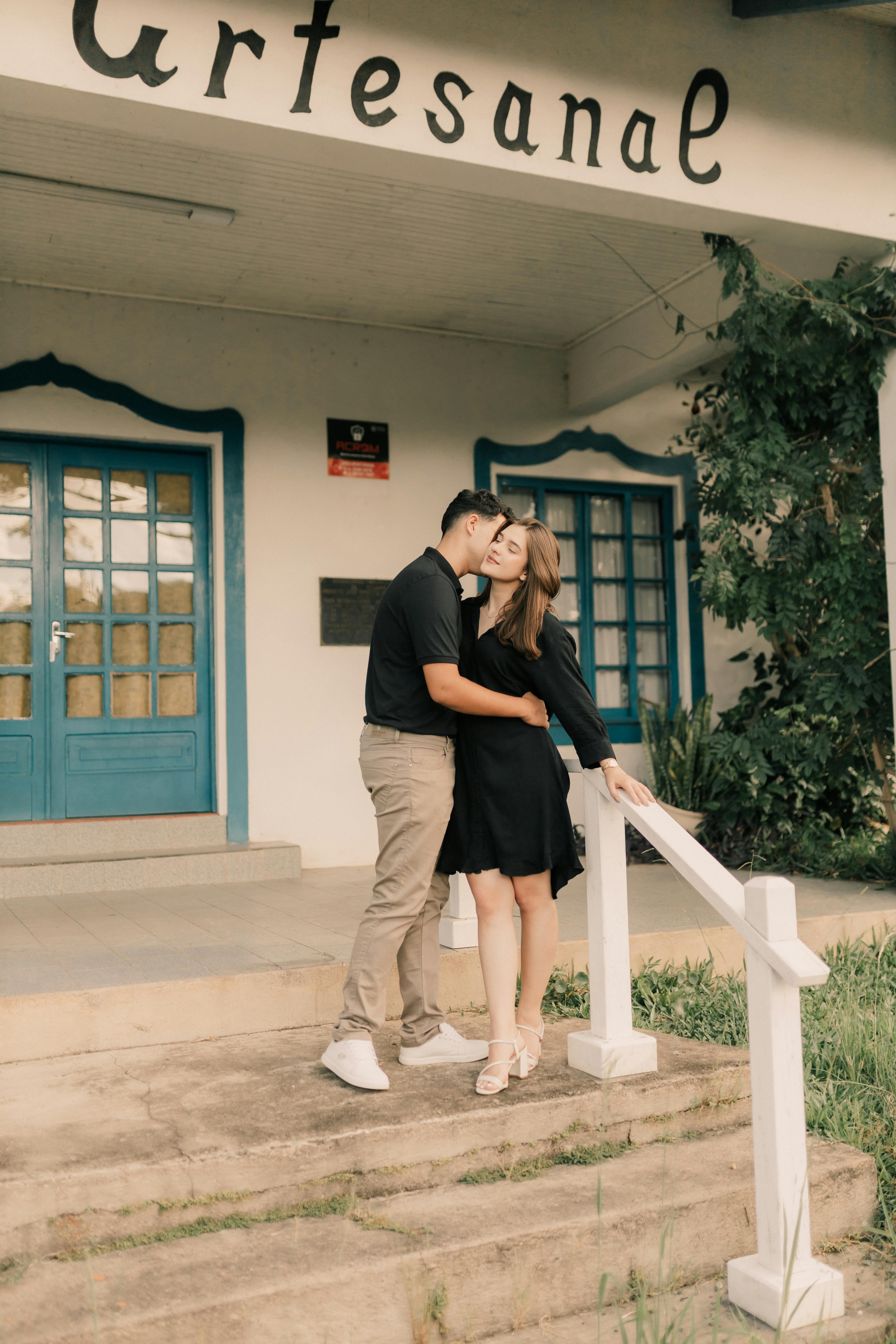 A couple shares a romantic moment on a building's porch under an 'artesanal' sign.