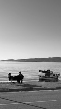 A serene lakeside view with a boat and two people on a bench, captured in black and white.