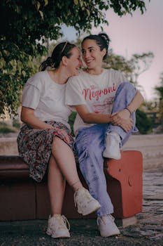 Two young women sitting together, laughing and enjoying a joyful moment outdoors on a bench.