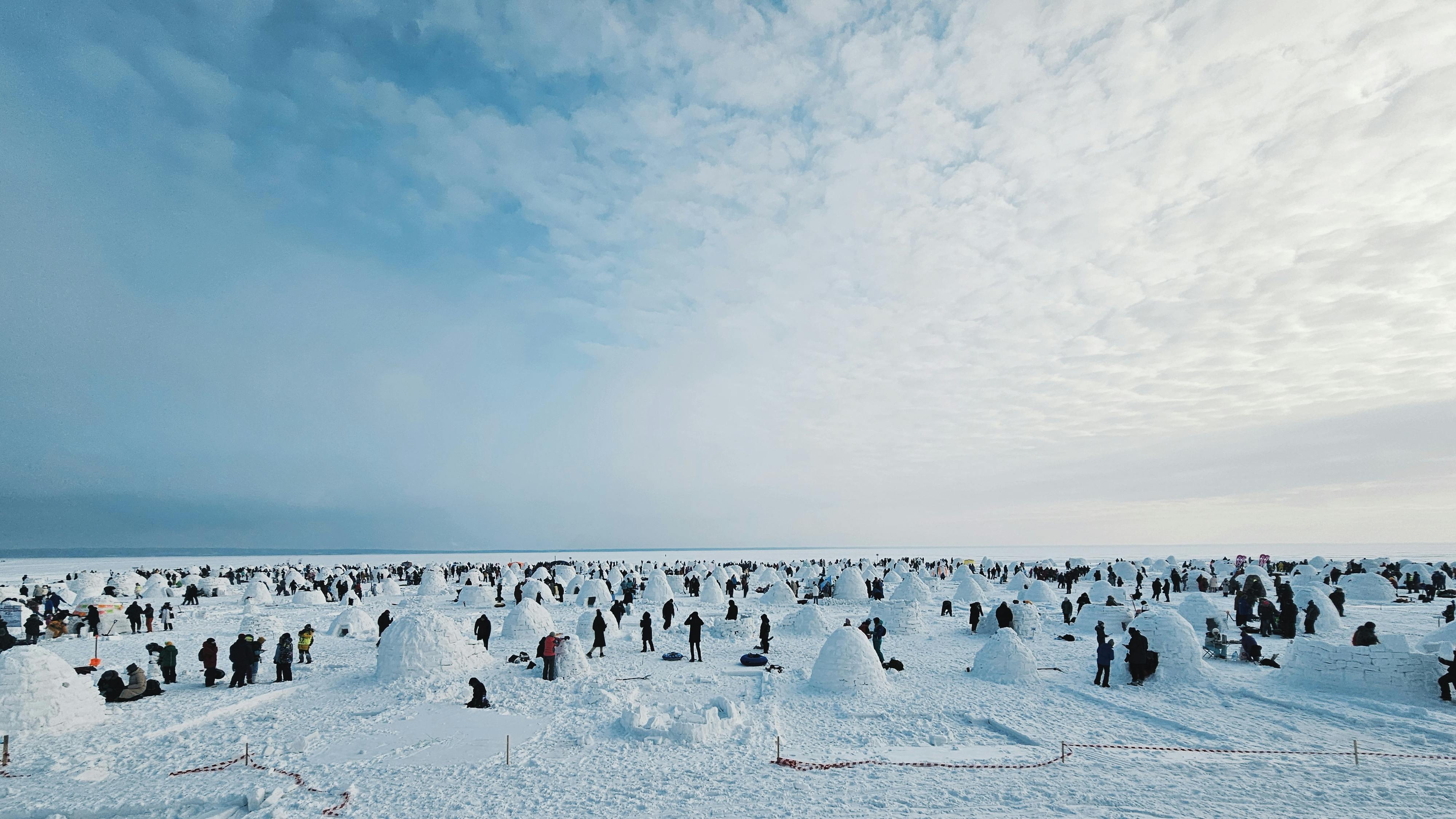 A vast snowy landscape with numerous igloos and people engaging in winter activities under a cloudy sky.
