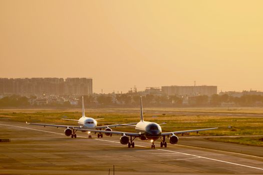 Two commercial airplanes taxiing at sunset with cityscape in background.
