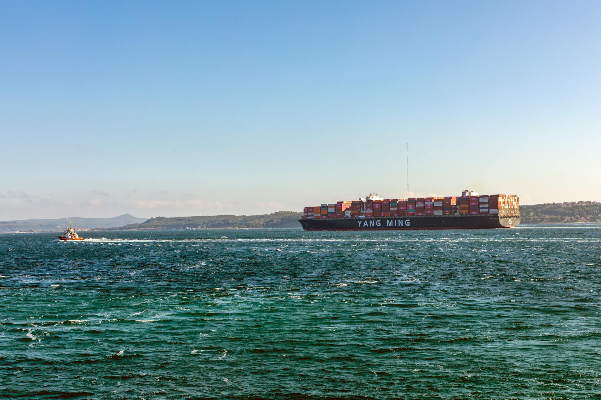 A Yang Ming cargo ship navigating the ocean with a small boat nearby under clear blue skies.