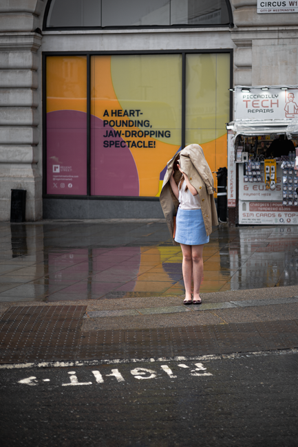 A woman shields herself from the rain with a coat near a colorful sign in London.