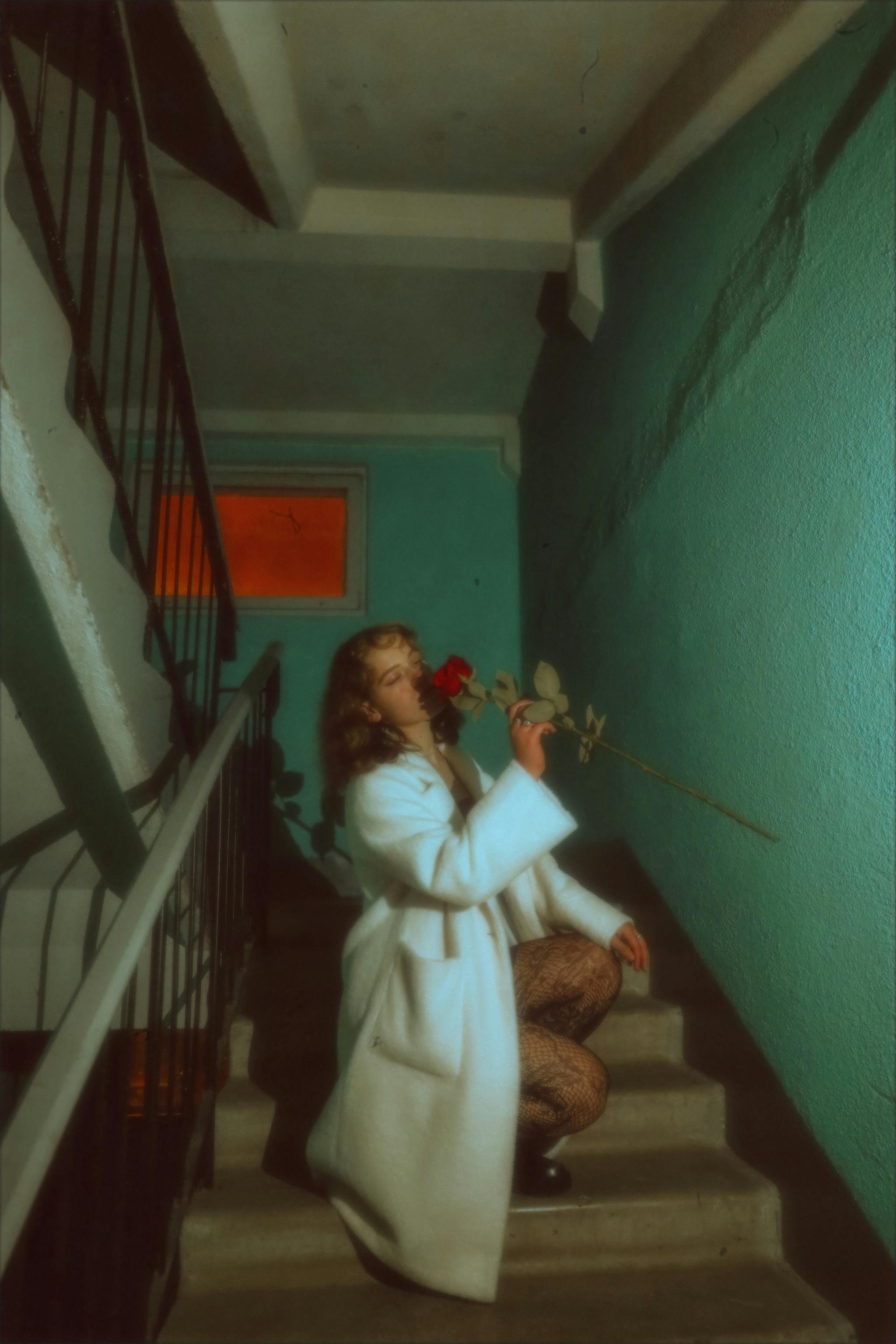 Stylish woman in white coat holds a red rose on dimly lit staircase.