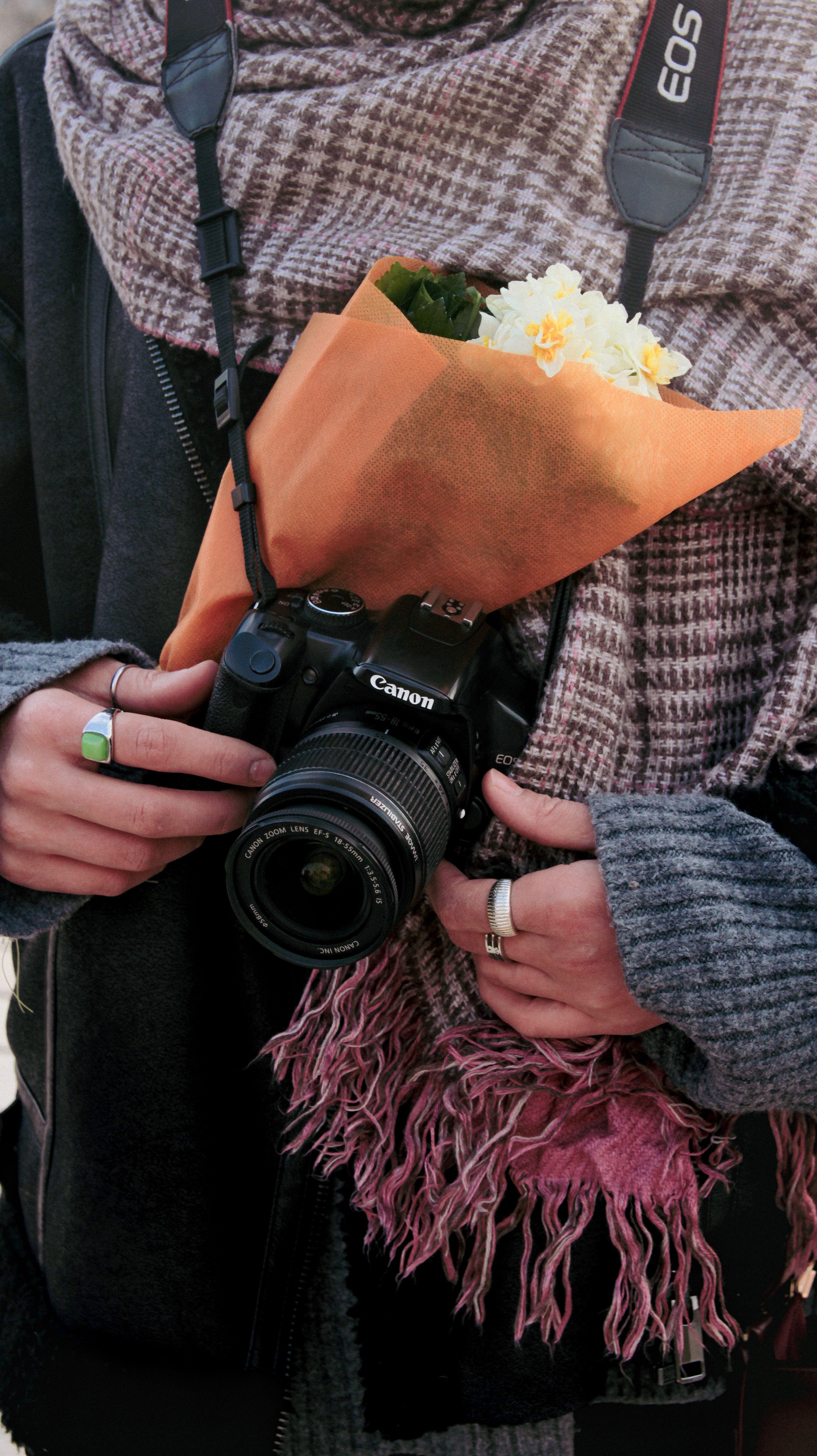 A photographer holds a DSLR and flowers, wrapped warmly in a scarf.