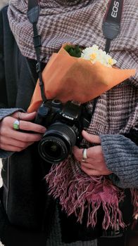 A photographer holds a DSLR and flowers, wrapped warmly in a scarf.