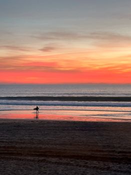 A lone surfer walks along a serene beach at sunset, under a vibrant sky.