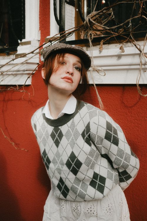 Free Stylish young woman in argyle sweater and newsboy cap posing against a red wall. Stock Photo