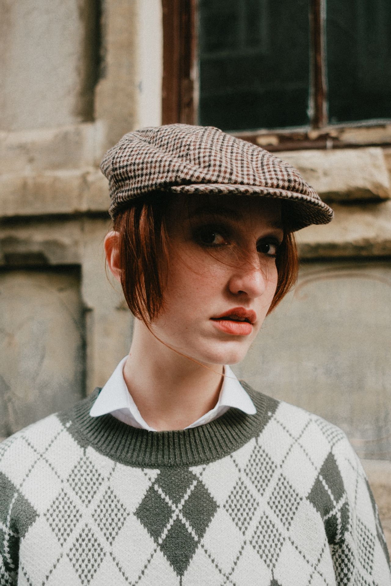 Woman wearing a gray and white argyle diamond check sweater over a white collared shirt with a check flat cap