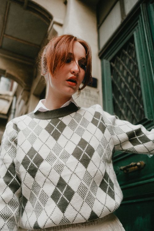 Free Young woman in cozy argyle sweater posing outside a classic building. Stock Photo