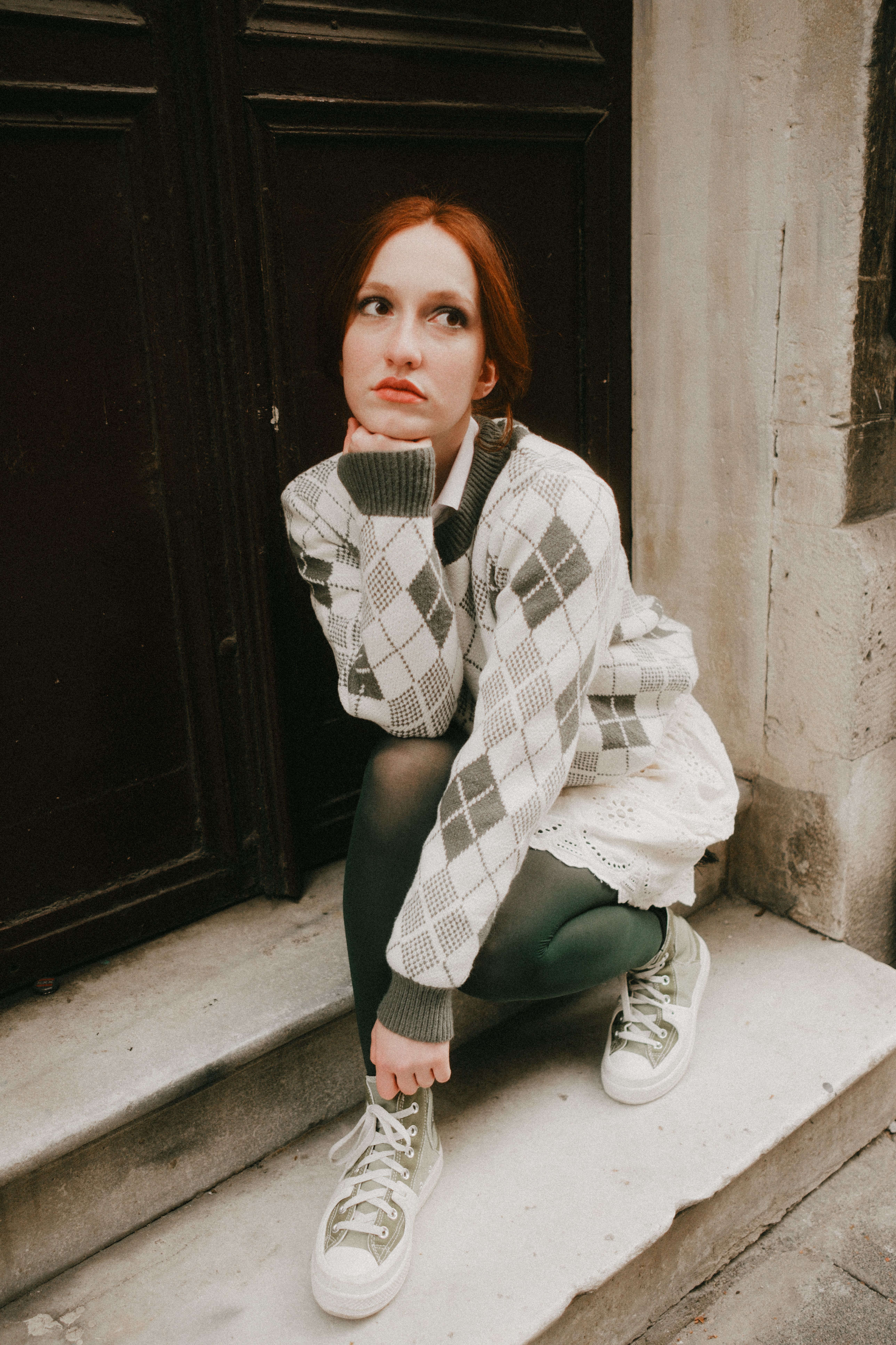 Red-haired woman in urban setting wearing a checkered sweater and sneakers, looking introspective.