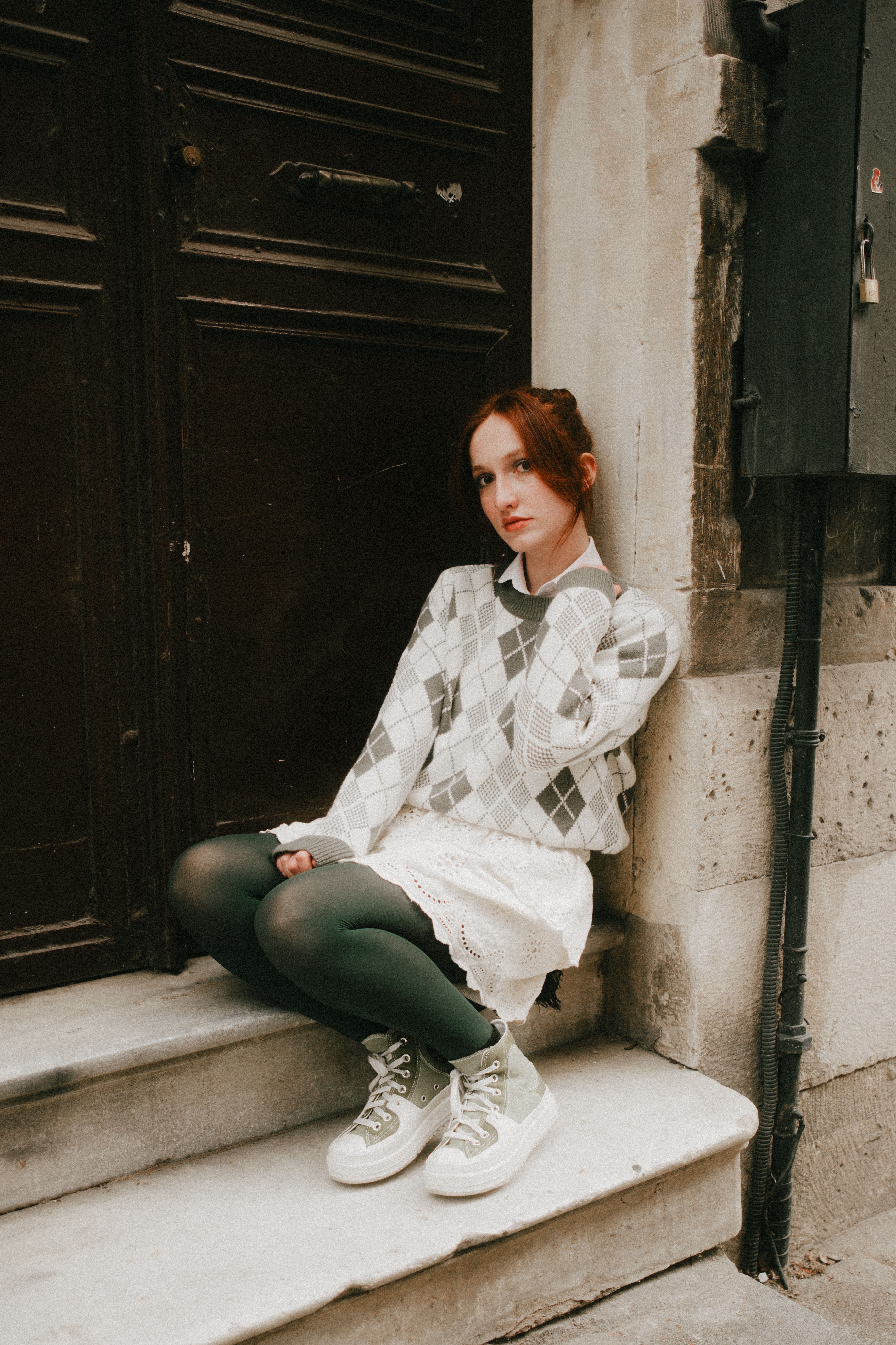 Young woman in stylish argyle sweater and sneakers sitting on steps outdoors.