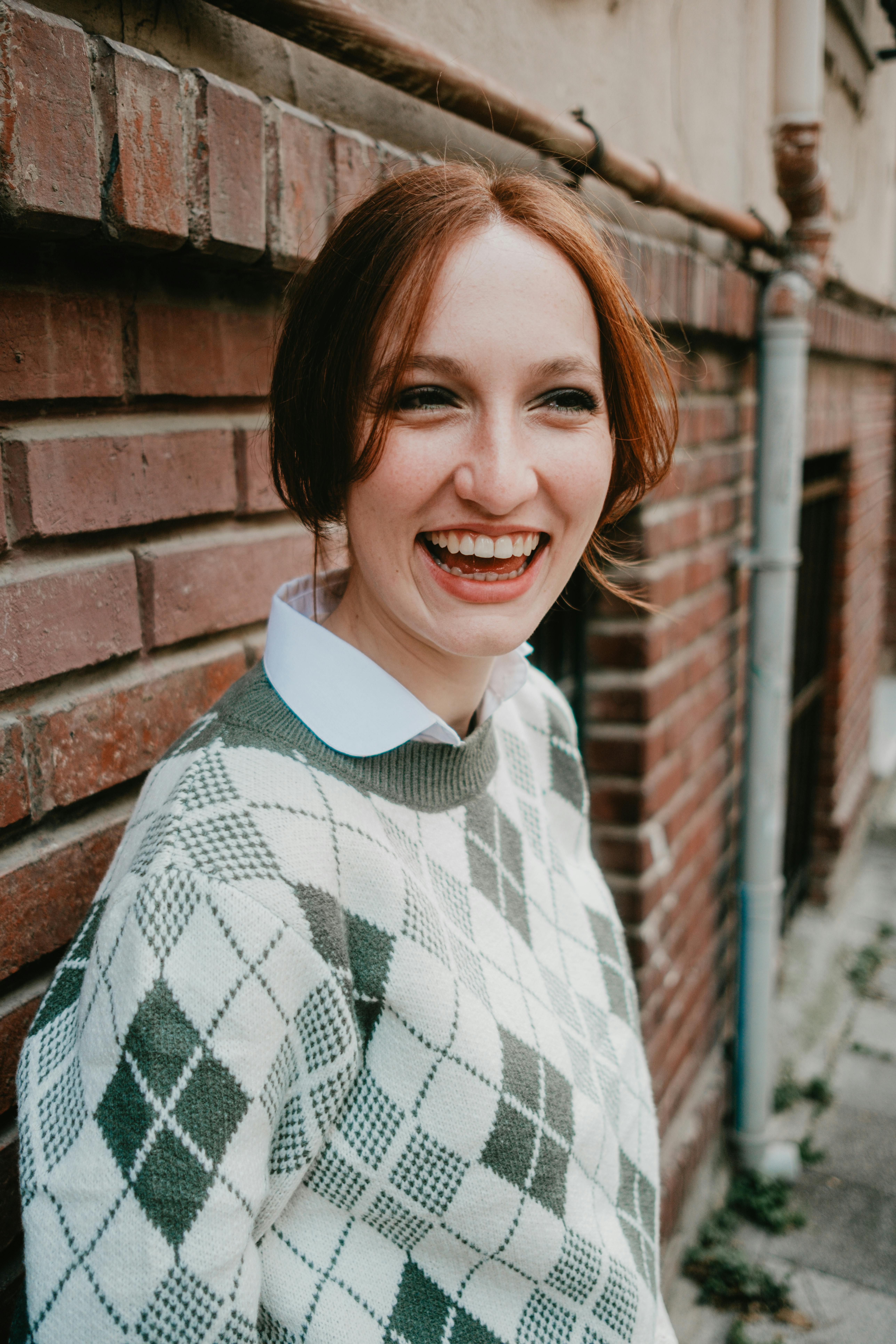 Young woman laughing, leaning on a rustic brick wall outdoors. Casual autumn fashion.