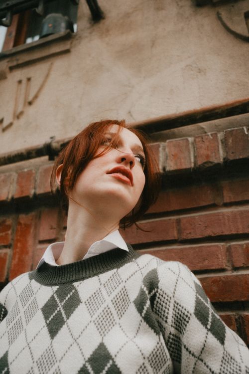 Free A thoughtful young woman in an argyle sweater stands against a brick wall. Stock Photo