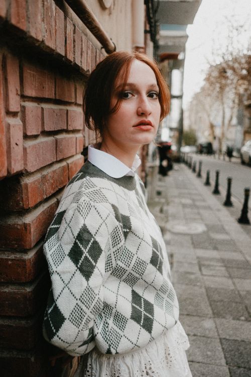 Free A young woman leans against a brick wall in an urban setting, wearing an argyle sweater. Stock Photo