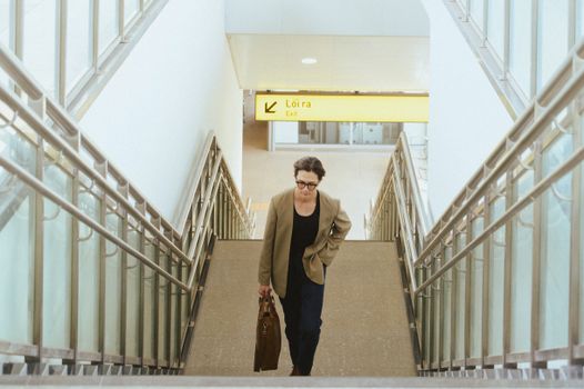 Businesswoman with briefcase climbing airport stairs towards exit sign marked 'Loira'.