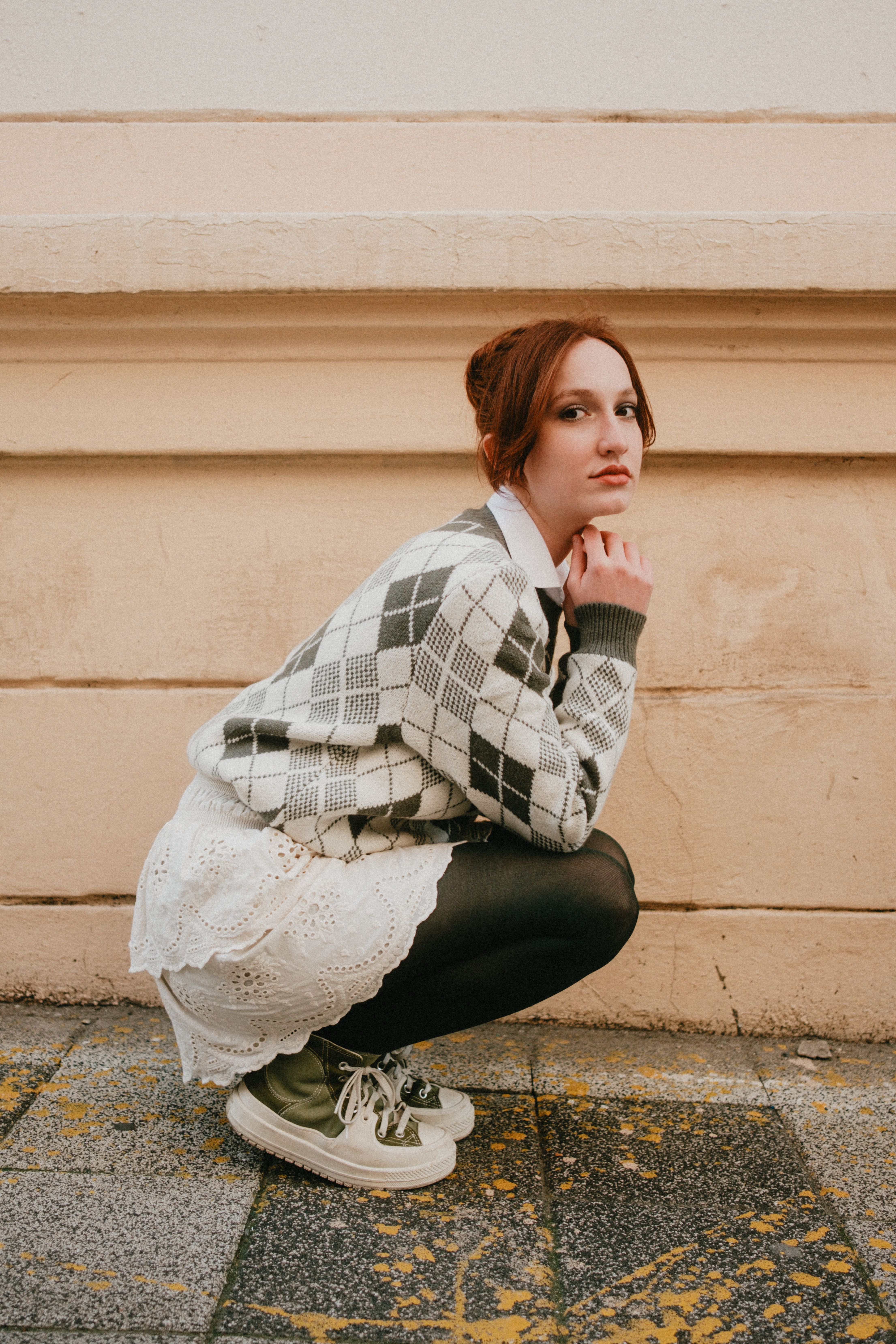 Young woman squatting in an argyle sweater and skirt on urban street.