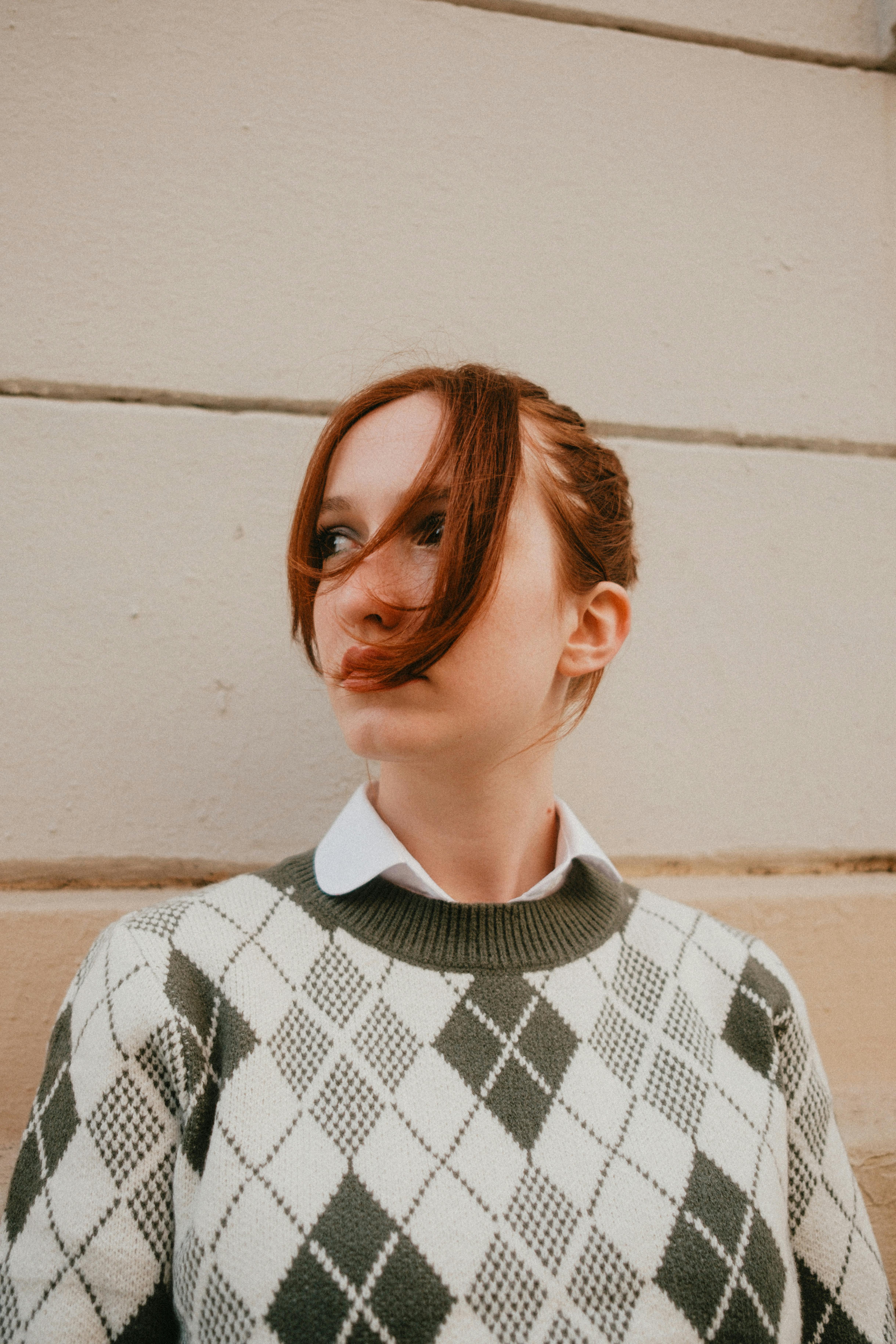 Stylish woman with red hair in argyle sweater against textured wall.