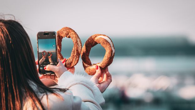 A person photographing two simit rings with a smartphone in an outdoor setting, creating a unique perspective.