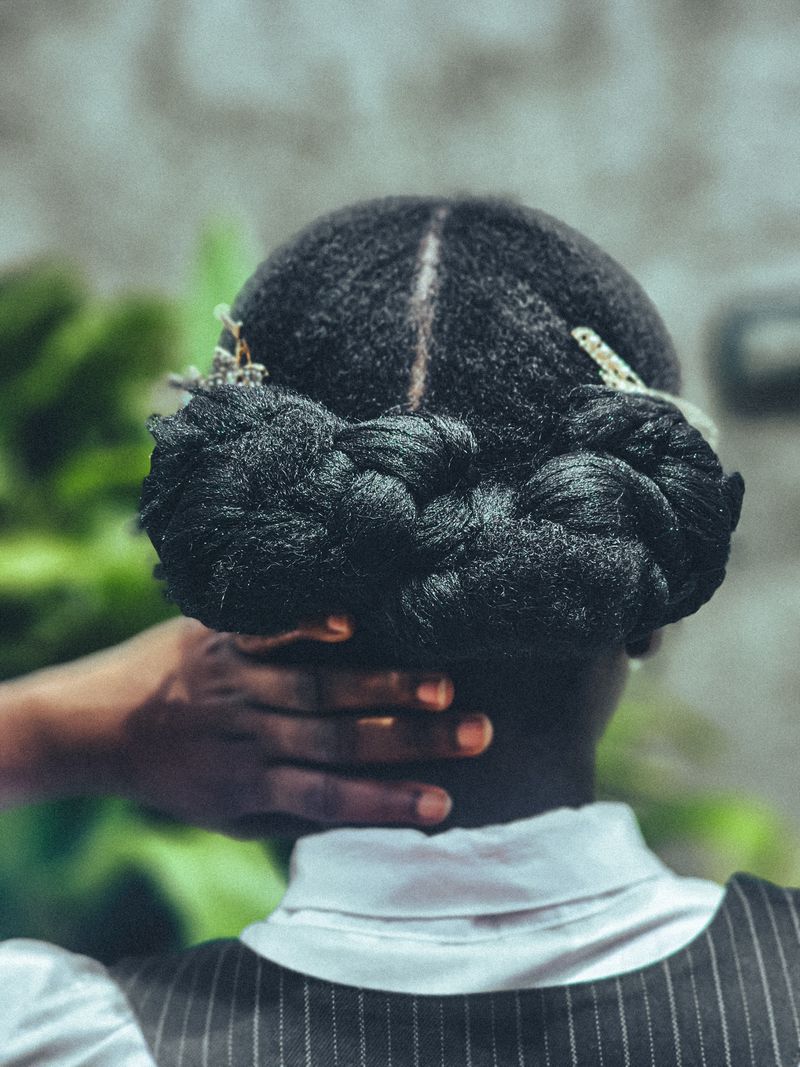 Close-up from behind of a Black woman with a braided updo and a center part on natural hair, one hand resting against the back of her head.