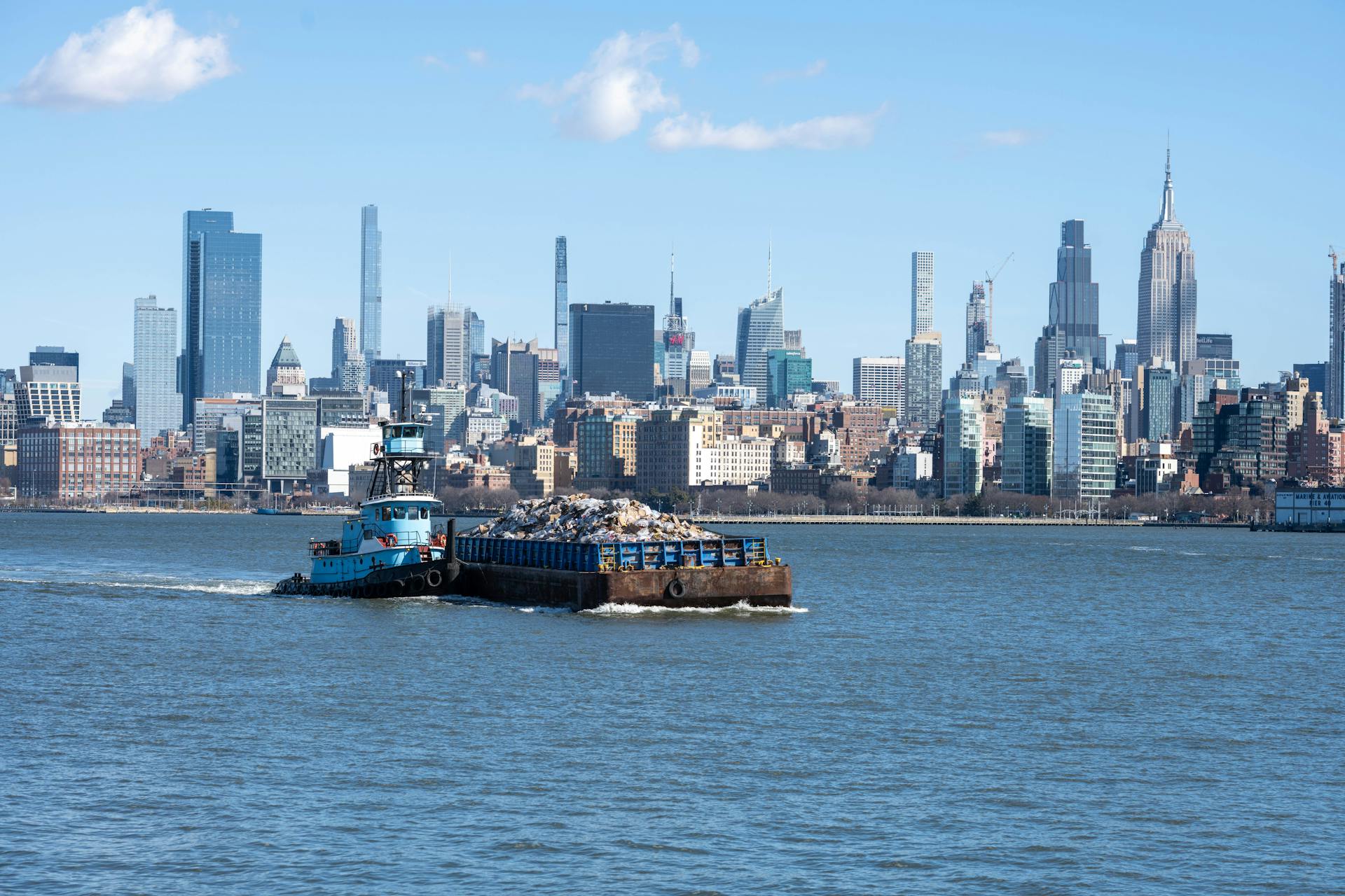 Tugboat towing barge on the Hudson River with iconic New York City skyline in the background.