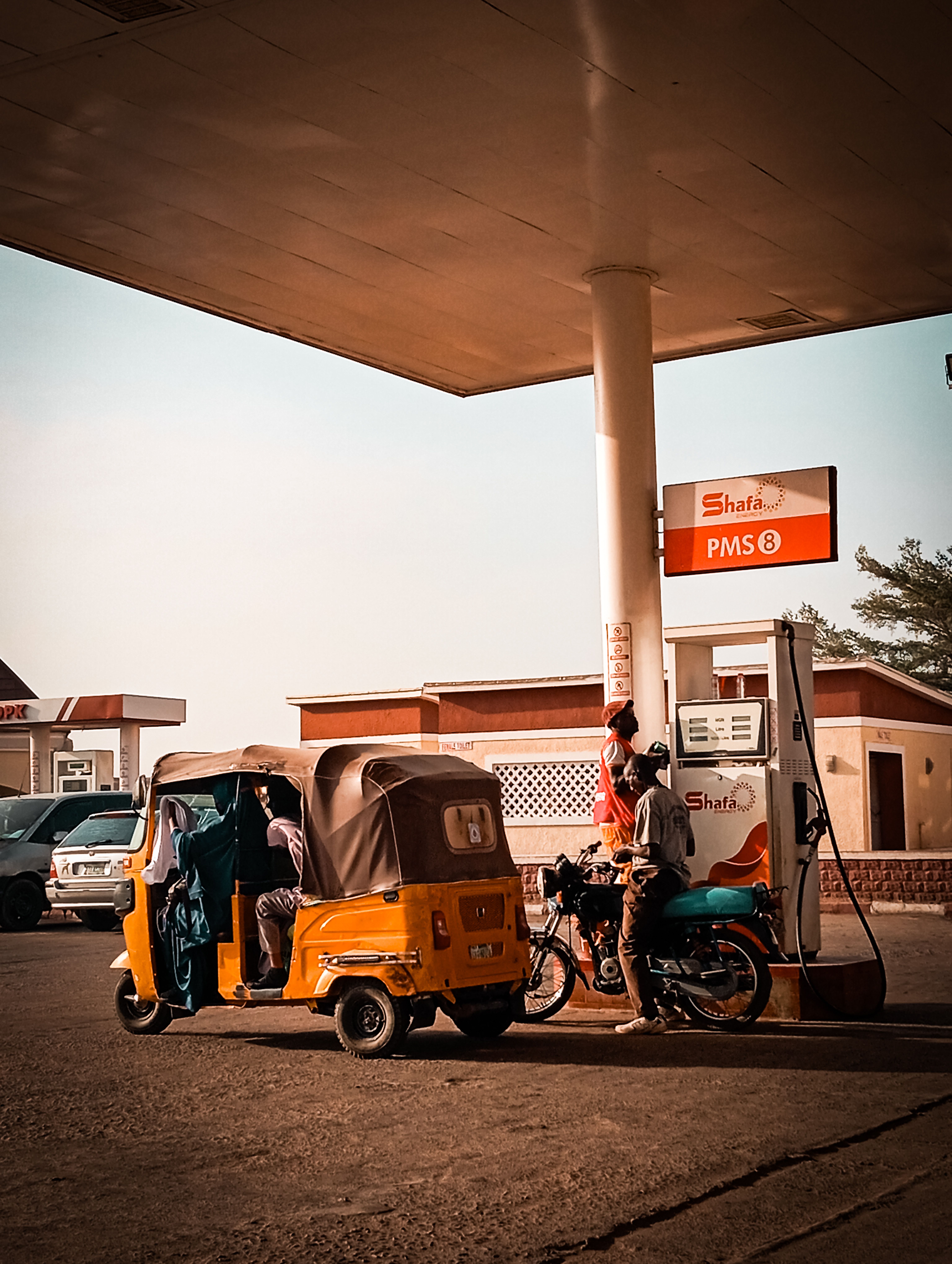 A vibrant scene at a gas station with a yellow rickshaw and motorbike refueling under a bright sky.