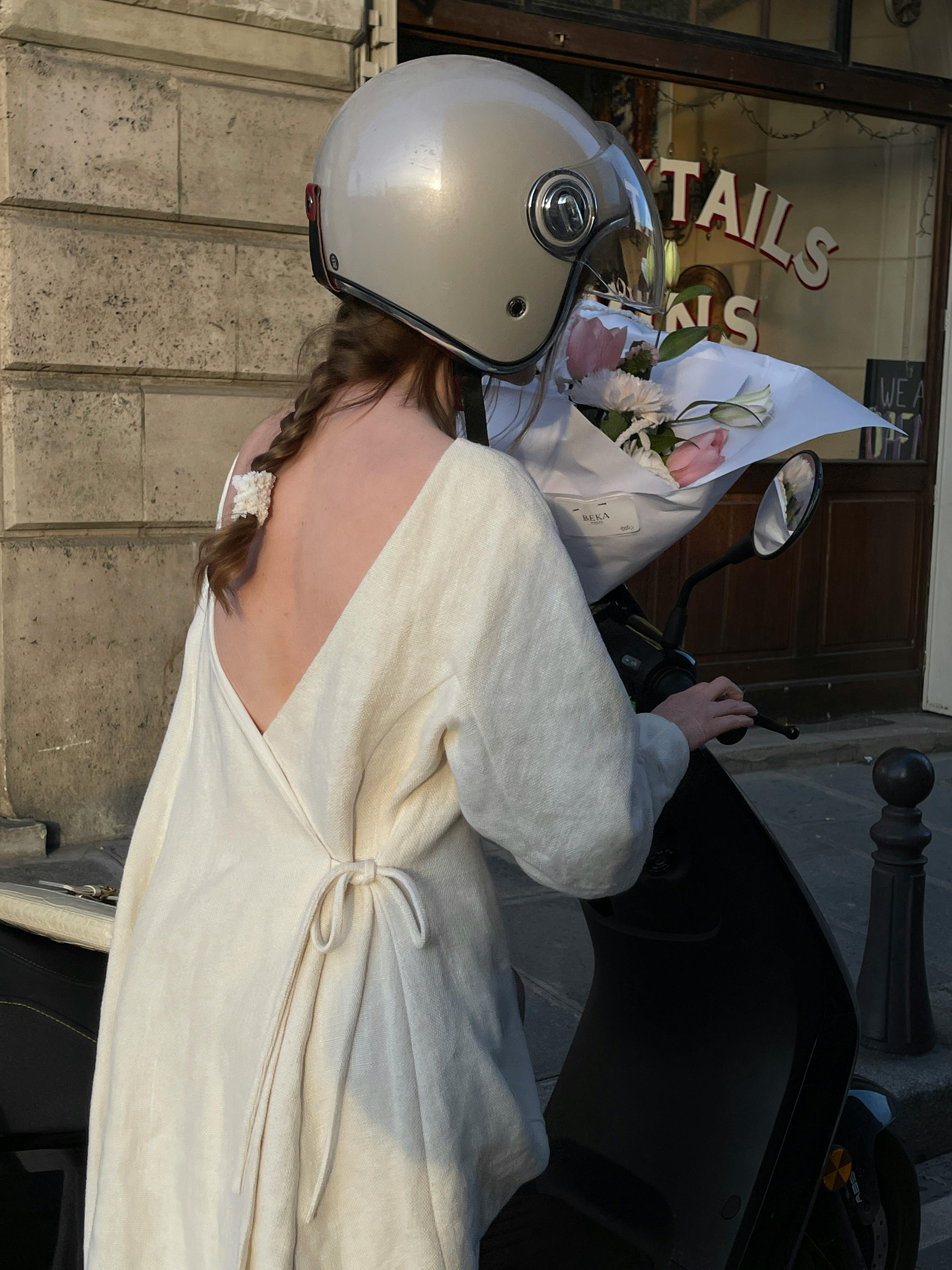 Woman in white dress and helmet, holding flowers on a scooter in a city.