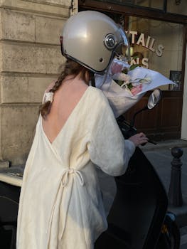Woman in white dress and helmet, holding flowers on a scooter in a city.
