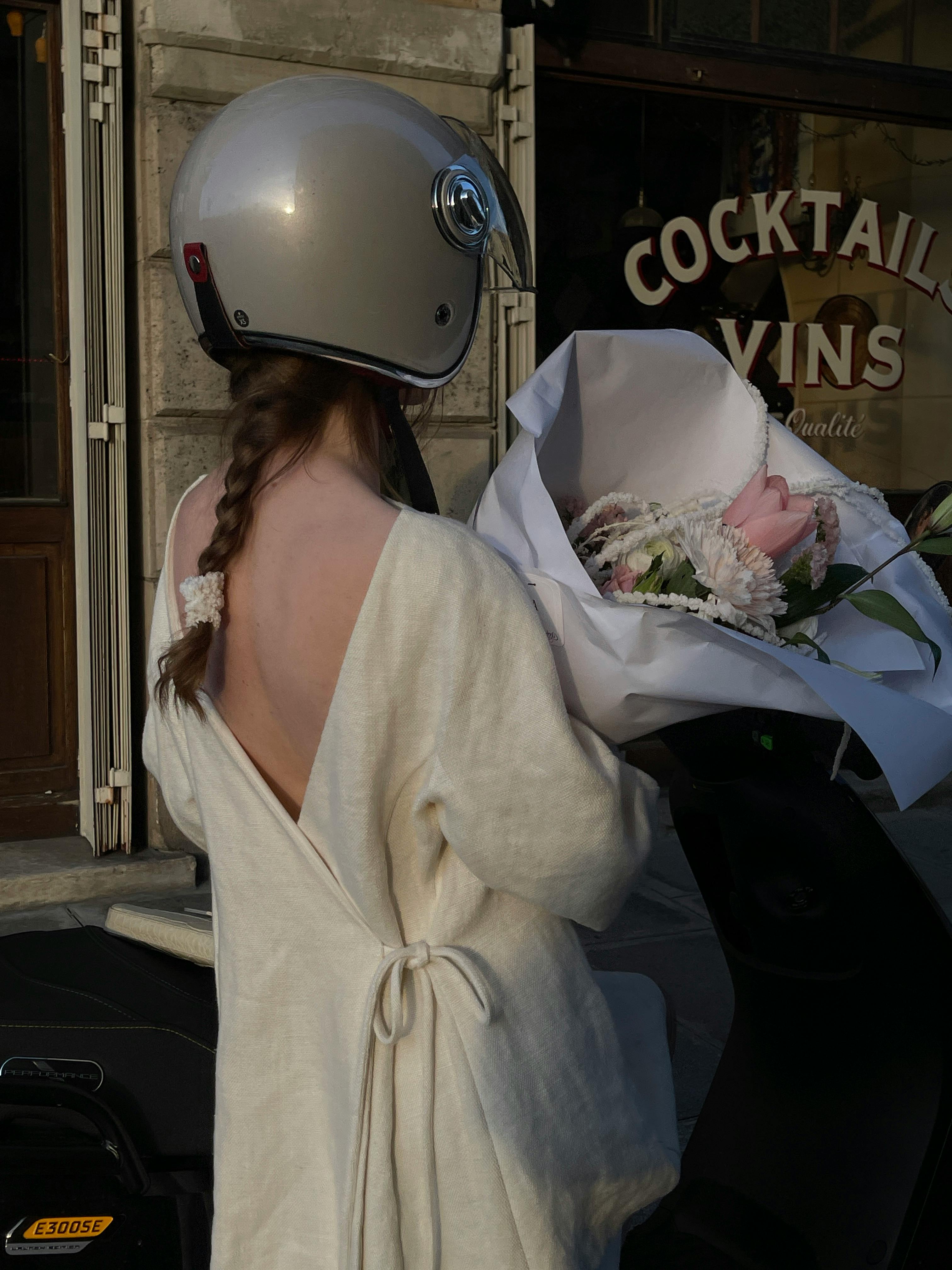 Woman in white dress with helmet holding flowers beside a scooter.