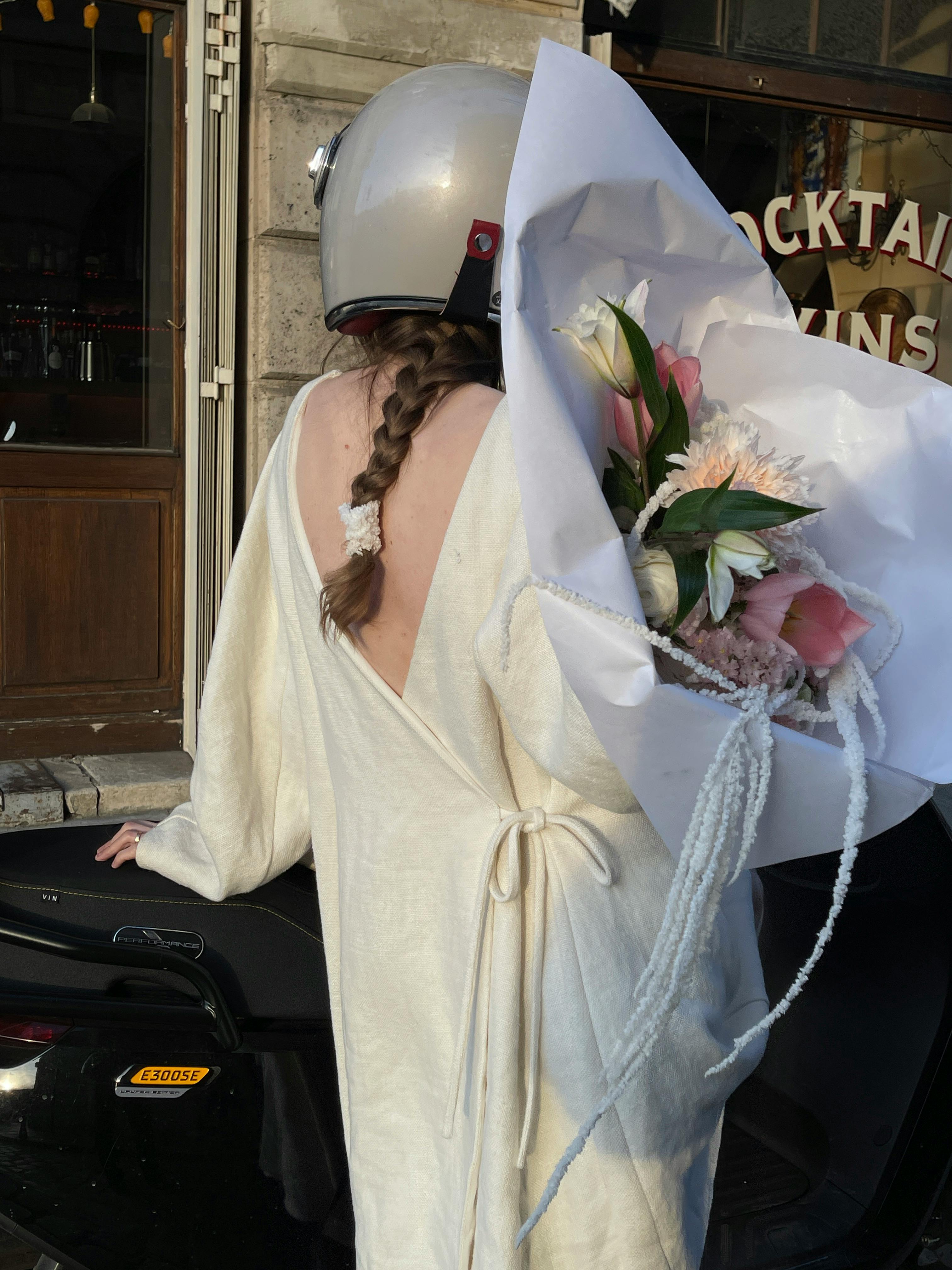 Woman in white robe with bouquet on scooter in city, exuding elegance and urban style.