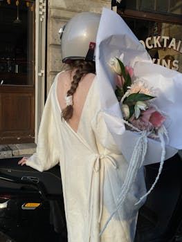 Woman in white robe with bouquet on scooter in city, exuding elegance and urban style.