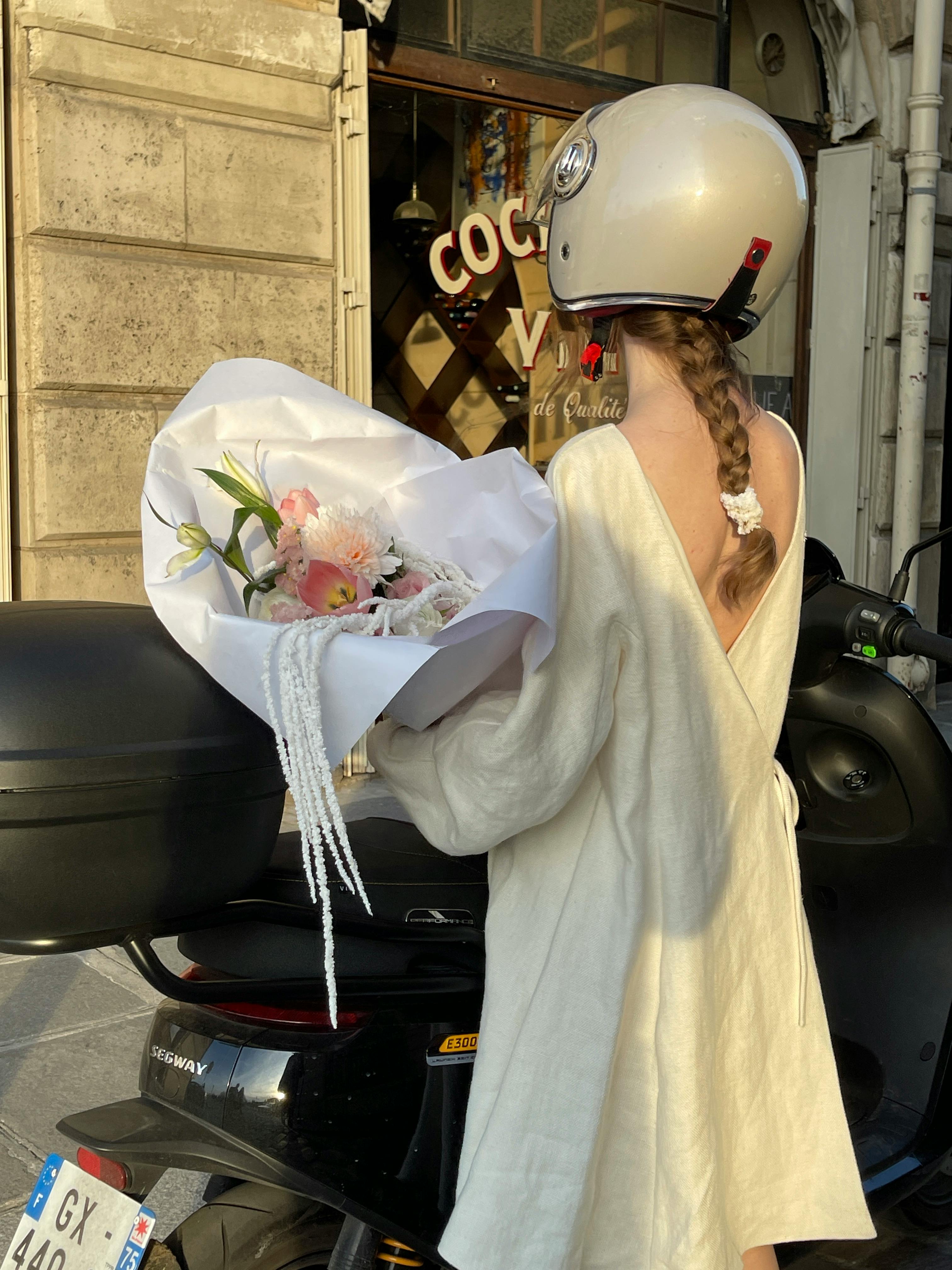 A woman with a helmet holds flowers next to a scooter, set in an urban street.