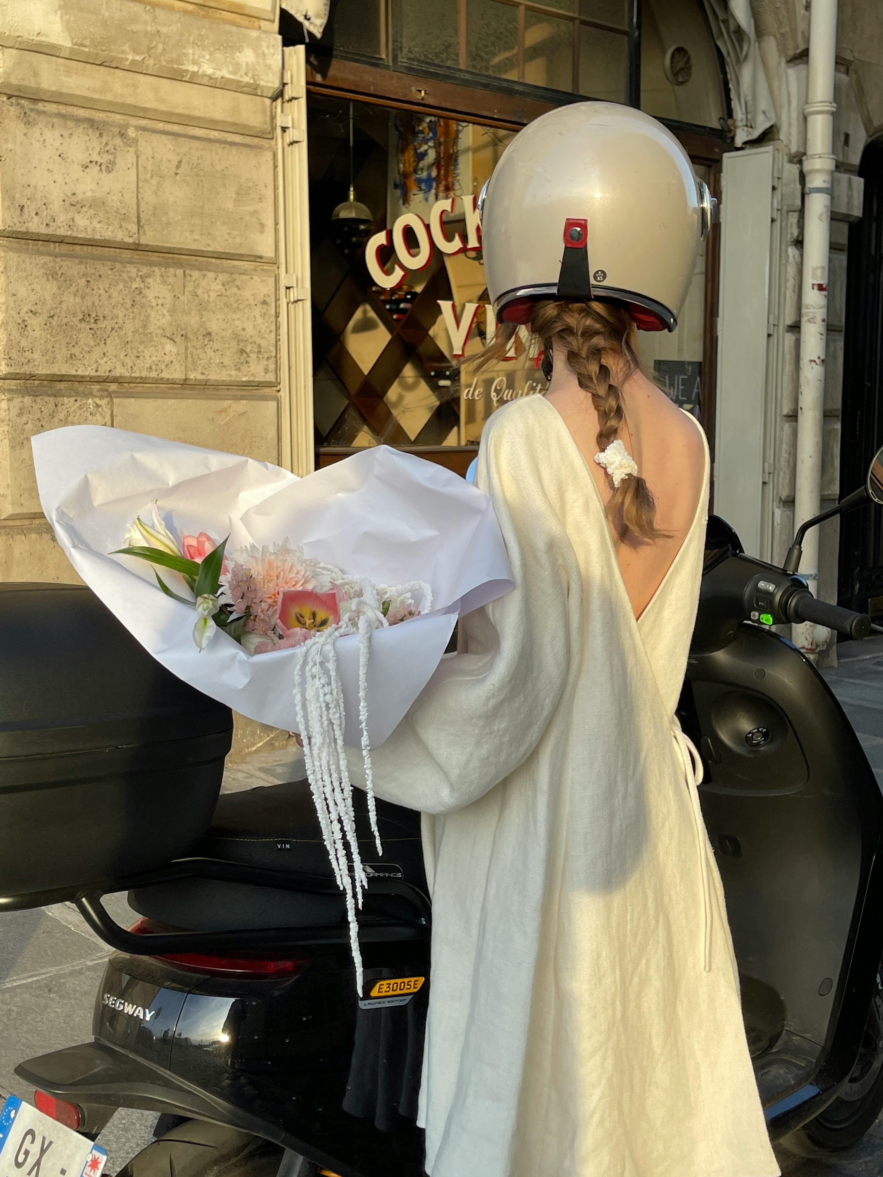 Woman in white dress holding flowers beside a scooter in urban surroundings.
